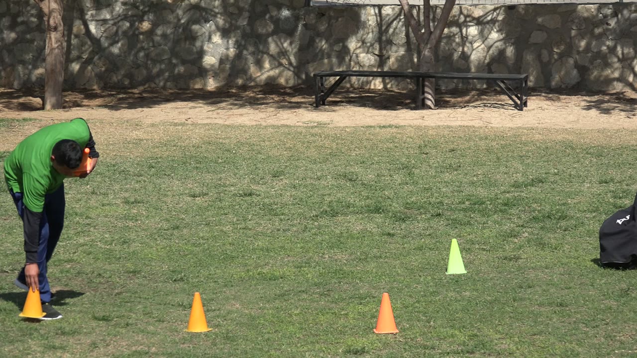A person in a green shirt bends beside an orange cone on a grassy training field in Los Cabos, Mexico, with more cones, trees, a stone wall, and a bench behind.