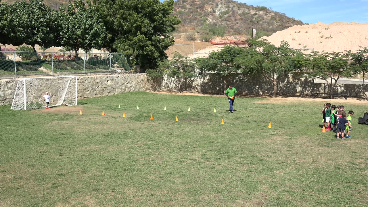 In Los Cabos, Mexico, the coach in a green shirt moves near the cone line while children remain clustered at the right edge of the field.