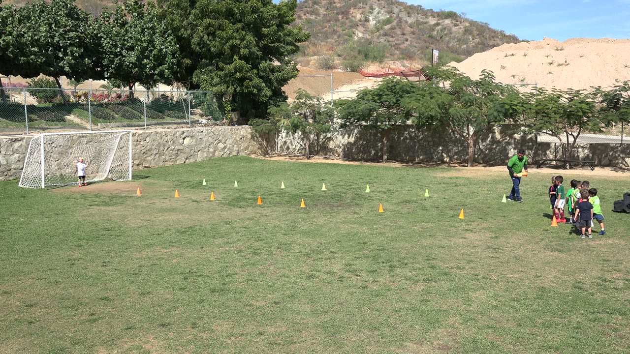 In Los Cabos, the coach leans toward the group of boys beside orange cones, with the white goal and hillside still visible behind them.