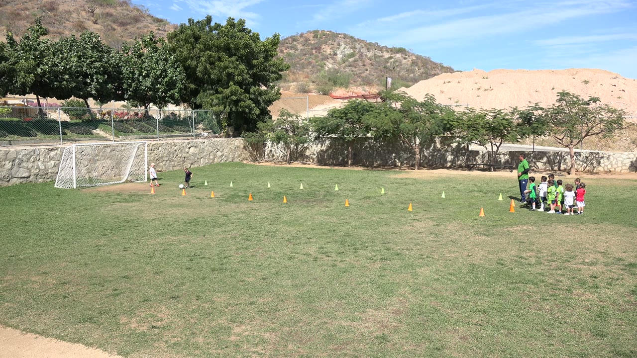 Wide frame of a Los Cabos park field where children gather near a coach on the right while one child stands near a white football goal on the left.