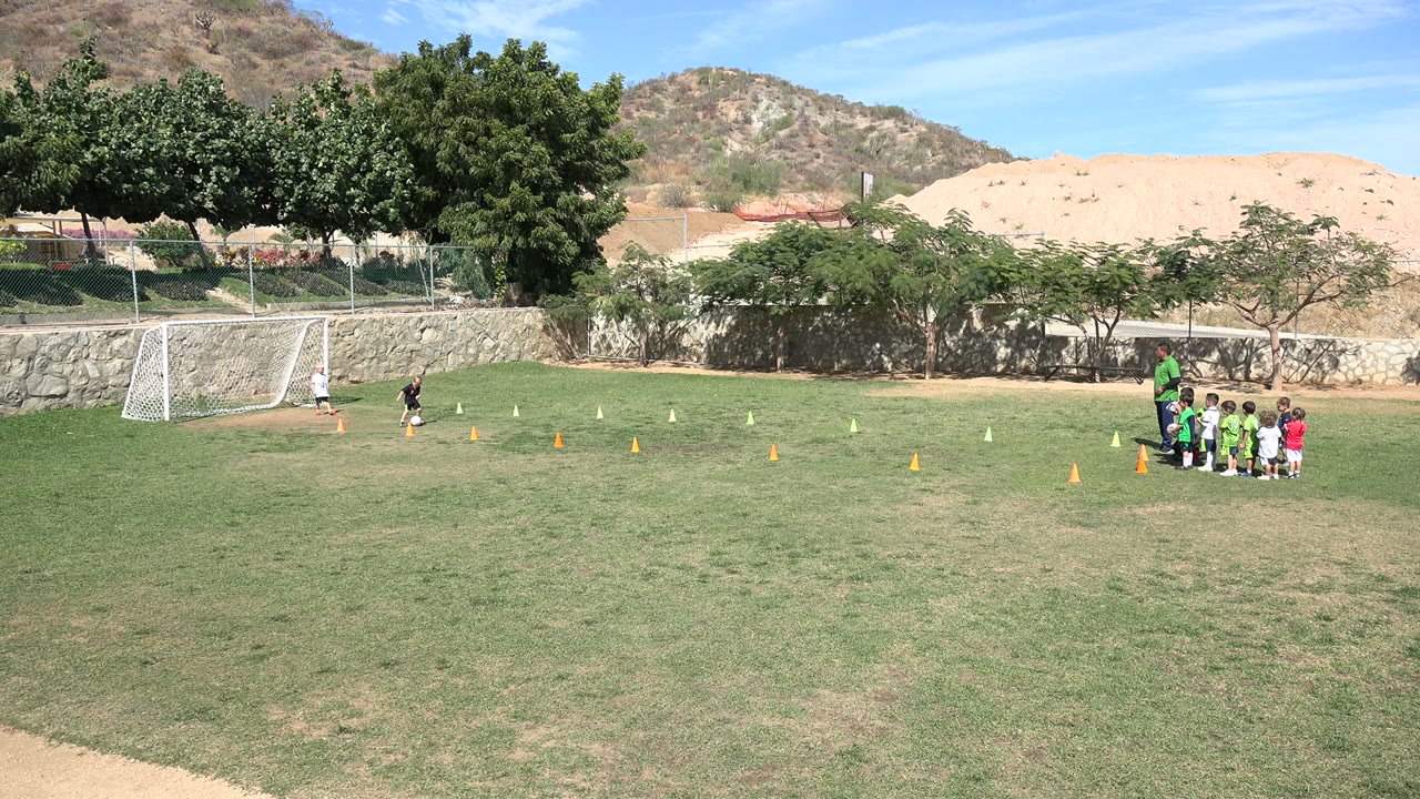 A child in dark clothing moves across the cone line in a Los Cabos park while the rest of the kids team waits in a cluster at the right side of the field.