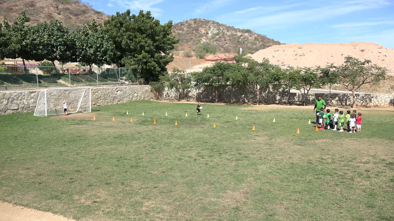 Wide Los Cabos soccer field with a coach standing beside young children near cones, a white goal at left, trees, stone wall, dry hills, and blue sky.