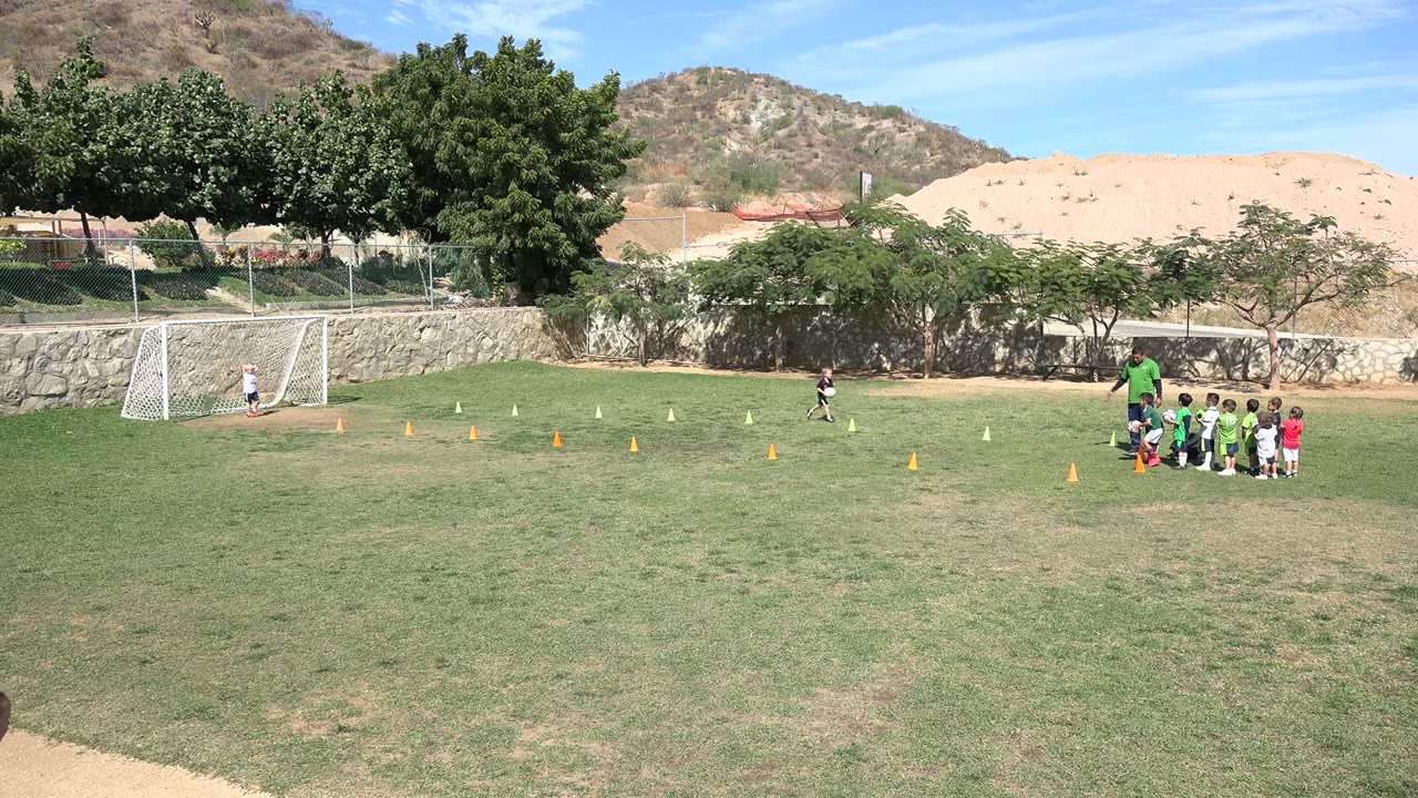A child moves across the Los Cabos grass field while the coach and children wait near training cones, with the goal, trees, wall, and hills behind them.