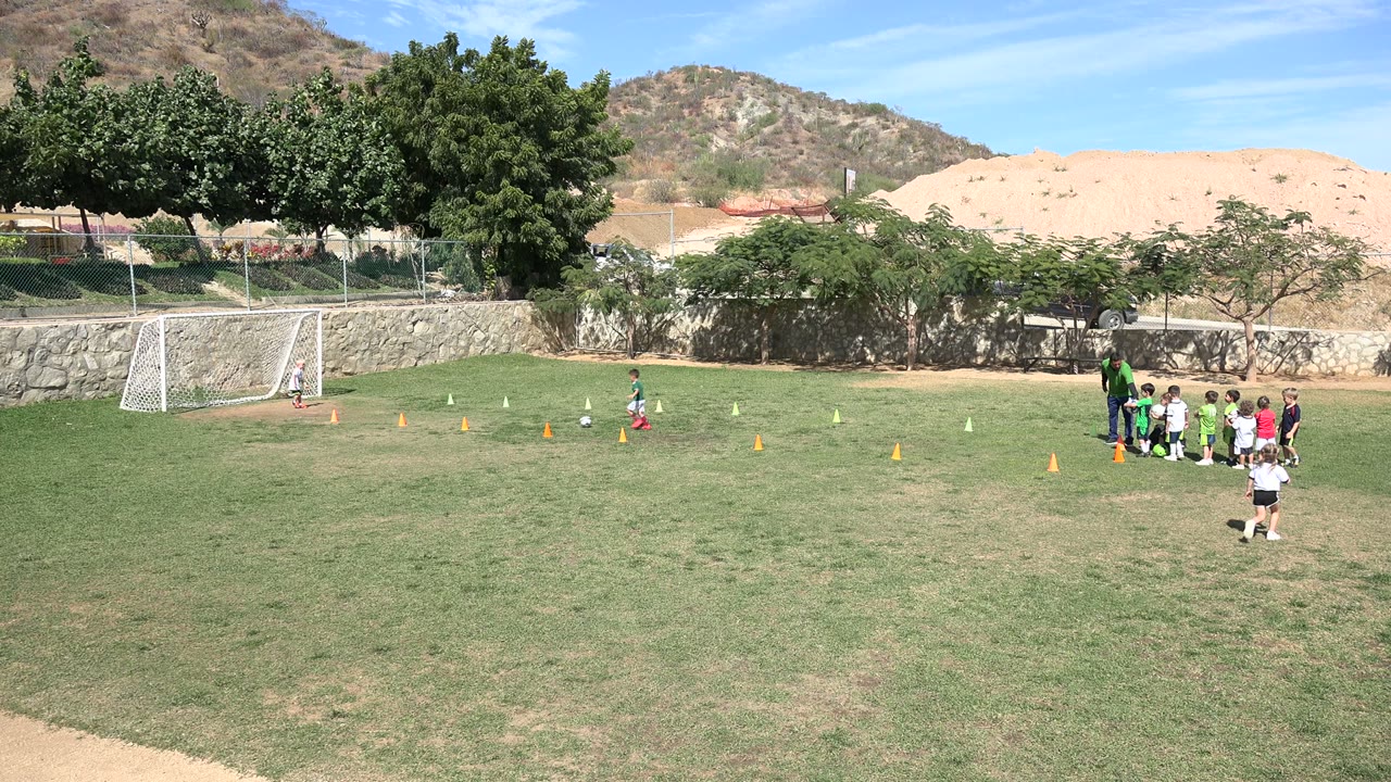 A child in soccer gear moves across the cone area on a sunny Los Cabos, Mexico field as a coach and several children wait near the right side of the frame.