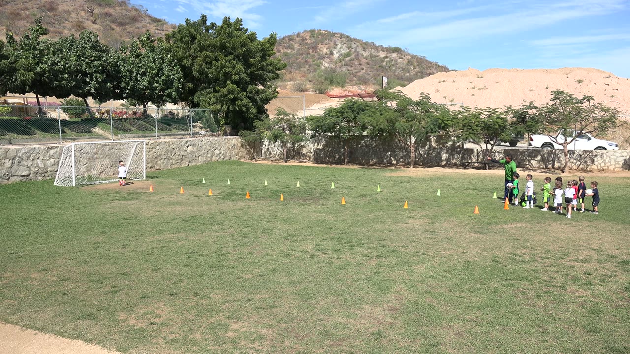 At 0:00 in Los Cabos, Mexico, children stand in a line behind a coach on a grass soccer field while a white goal, cones, trees, and dry hills sit behind them.