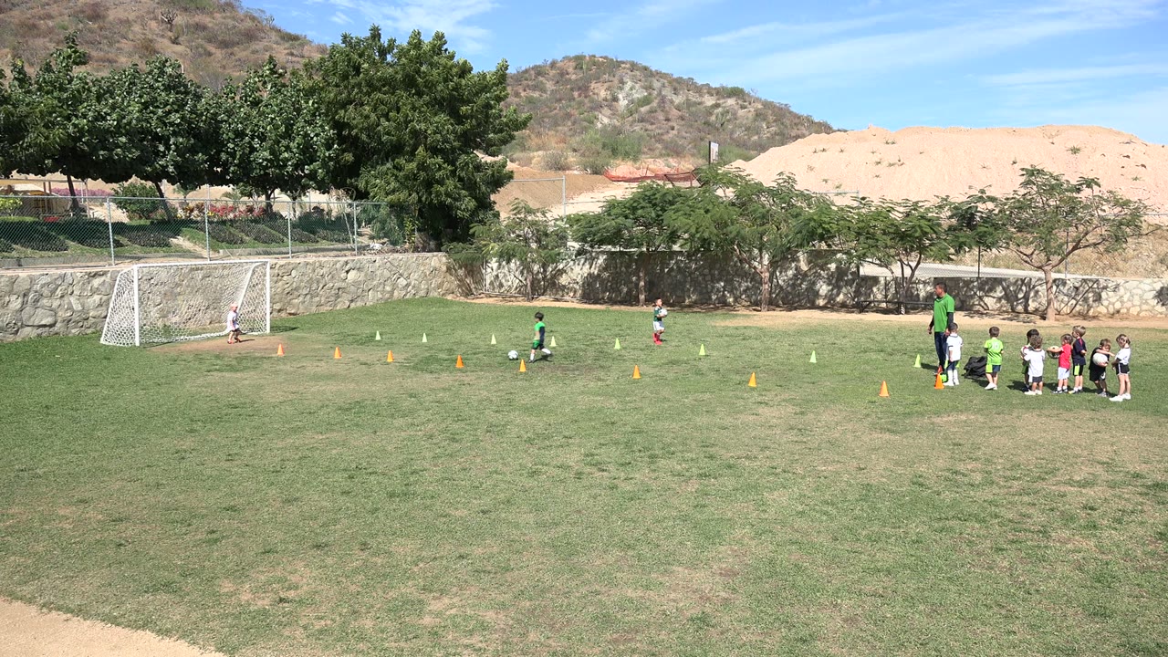 A young player in green works through the football drill in Los Cabos, Mexico, with cones across the grass and a line of children gathered near the adult coach.