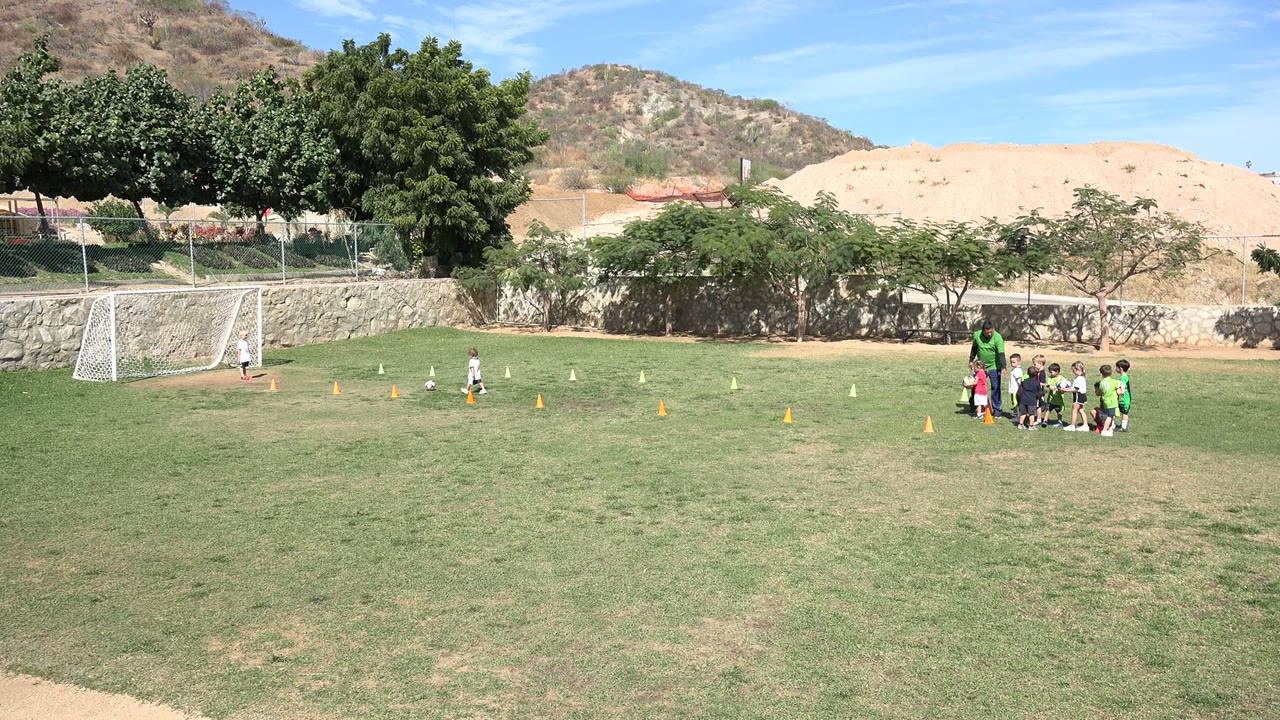 Wide frame of a child moving across the Los Cabos soccer field near the cones while the coach and team remain grouped to the right of the goal area.