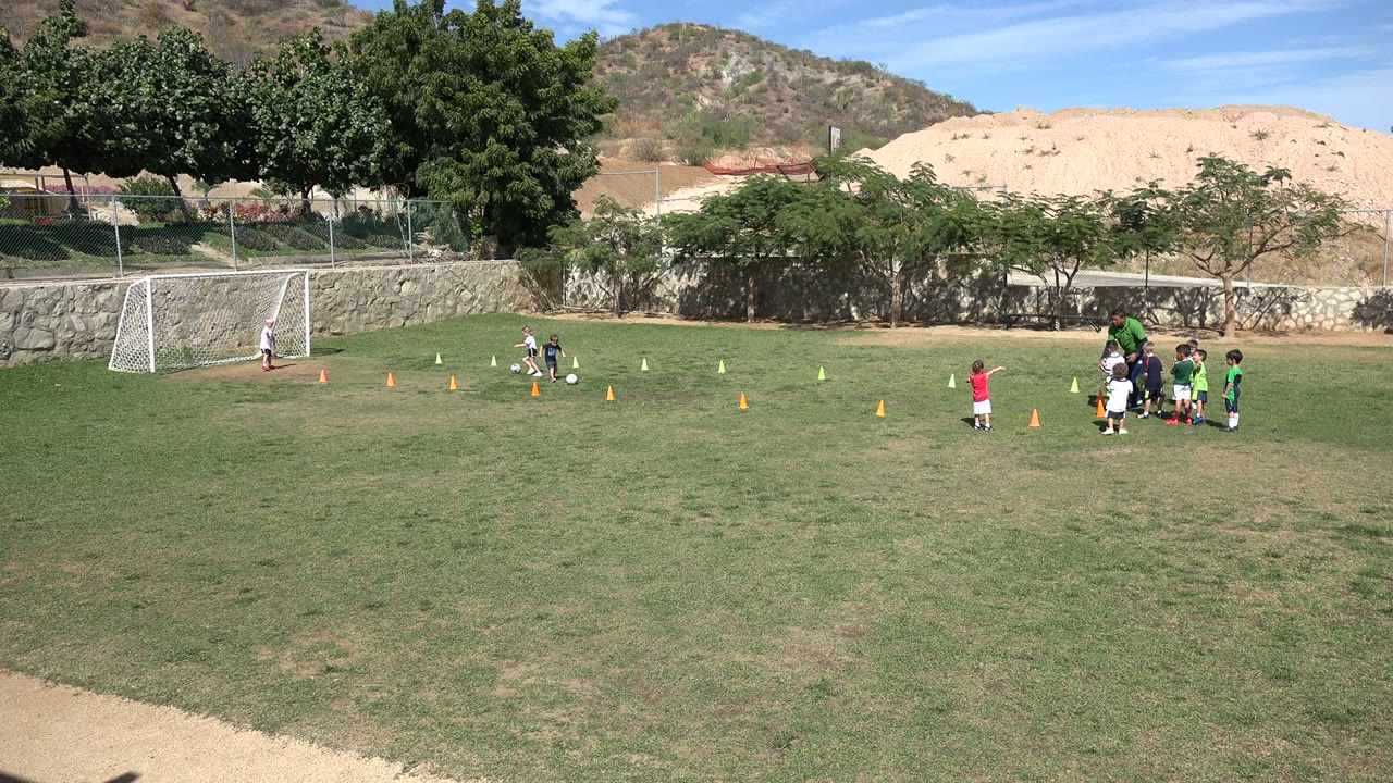 A child moves with a soccer ball near the left side of the drill area in Los Cabos, Mexico, while other children gather with an adult on the right.