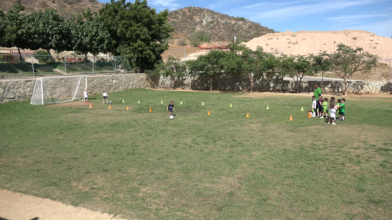 A child moves with a soccer ball near the center of the field in Los Cabos, Mexico, while other children gather at the right beside rows of training cones.