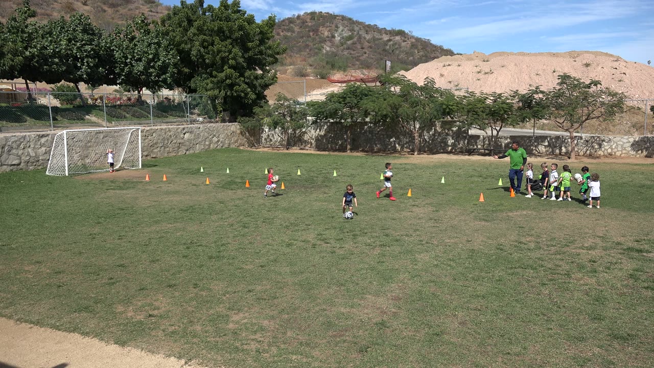 Children move between cones on the Los Cabos soccer field as the coach stands by a grouped line of players, with a goal at left and trees along the back wall.