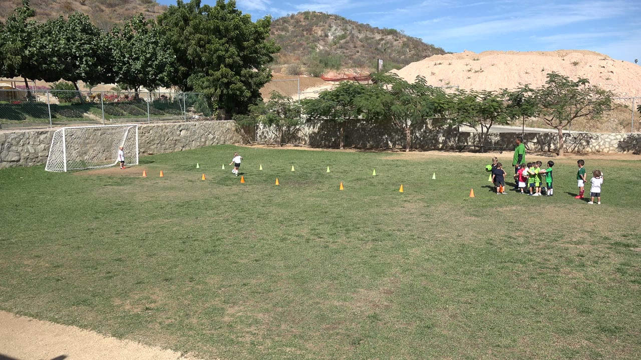 A child in a white shirt moves near the cone drill while the youth soccer team and coaches remain grouped on the right side of the Los Cabos field.