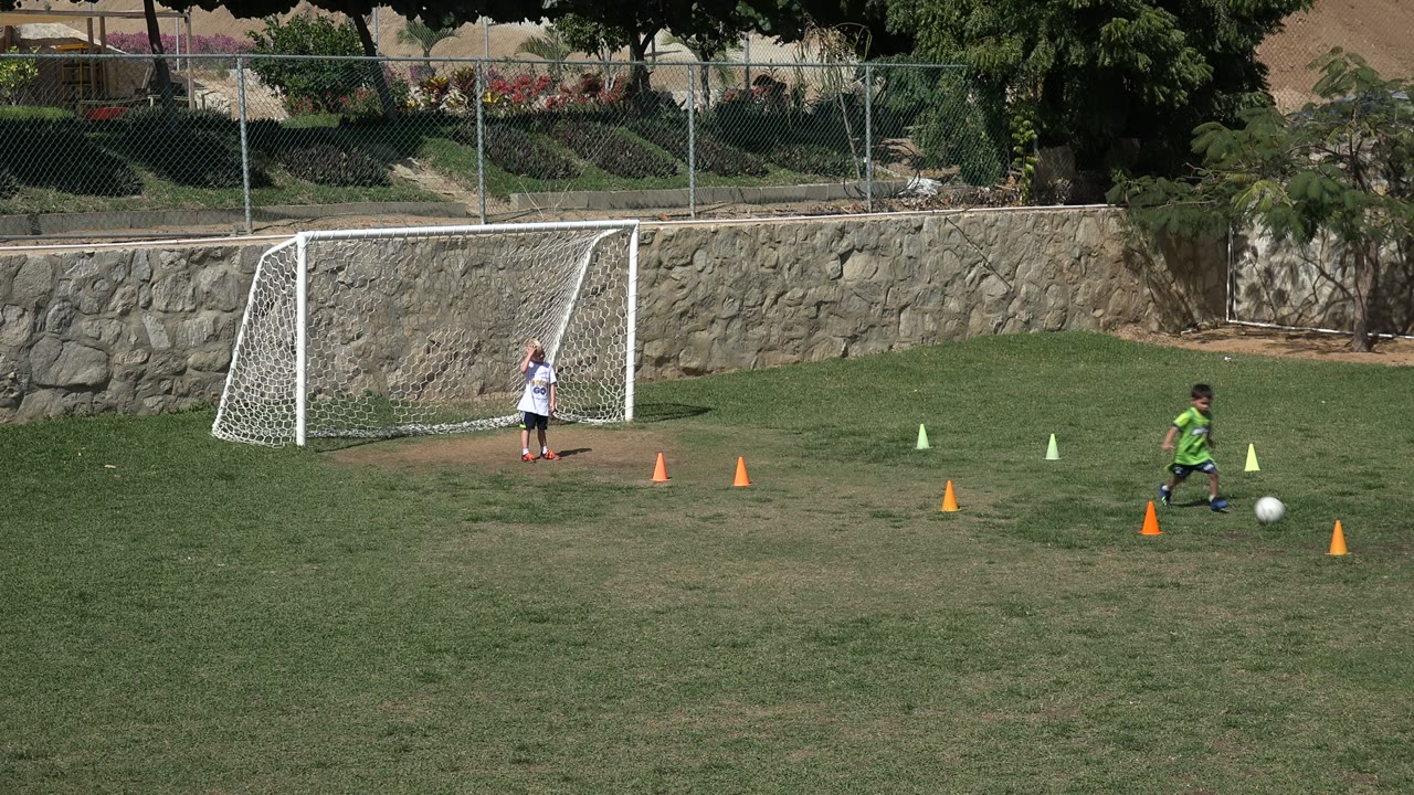 A child in green continues across the Los Cabos soccer field with the ball near orange cones as another child remains near the goal in the background.
