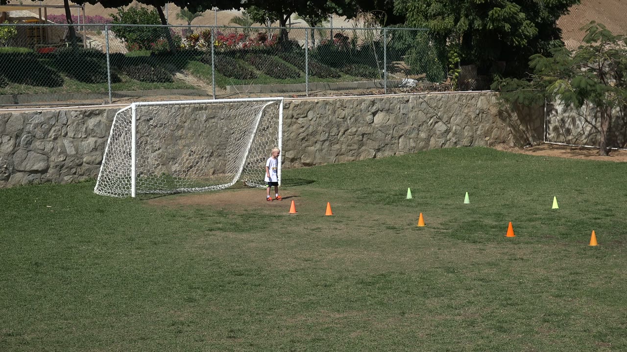 A child stands near a white soccer goal on a Los Cabos grass field while orange and pale green cones mark a drill area in the sun.
