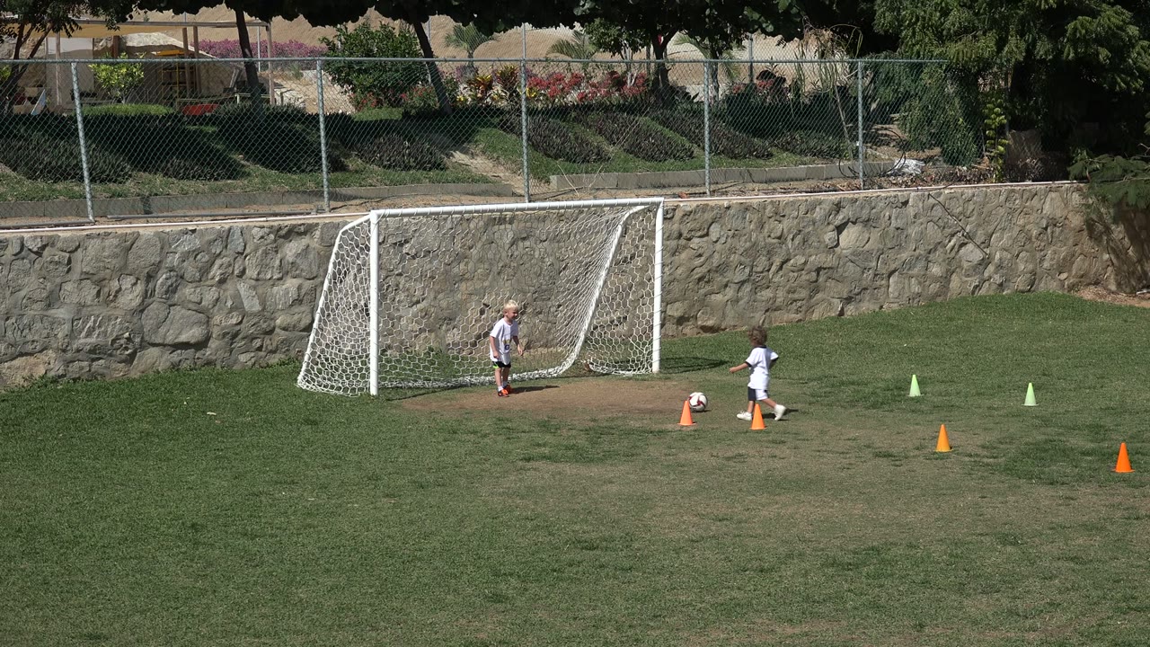 The striker moves the ball toward the small goal in Los Cabos, Mexico, while the other boy stands near the net and practice cones mark the grass.