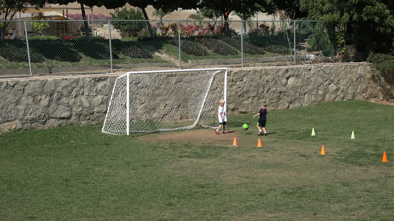A child in a white shirt and a child in dark clothing move near a green soccer ball beside a white goal on the Los Cabos grass field.