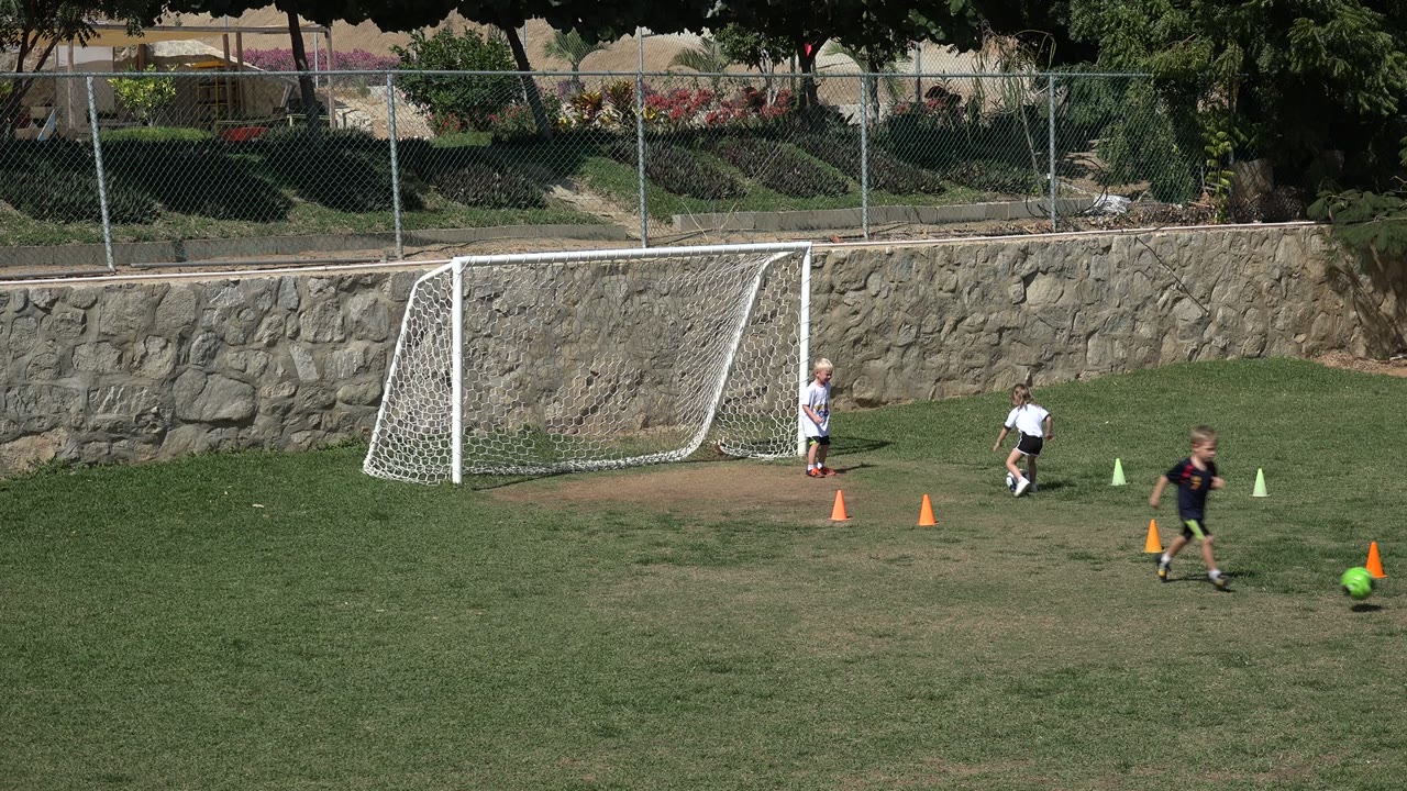 In Los Cabos, Mexico, children are spread across a grass football field near a white goal, orange cones, a stone wall, and a chain-link fence.