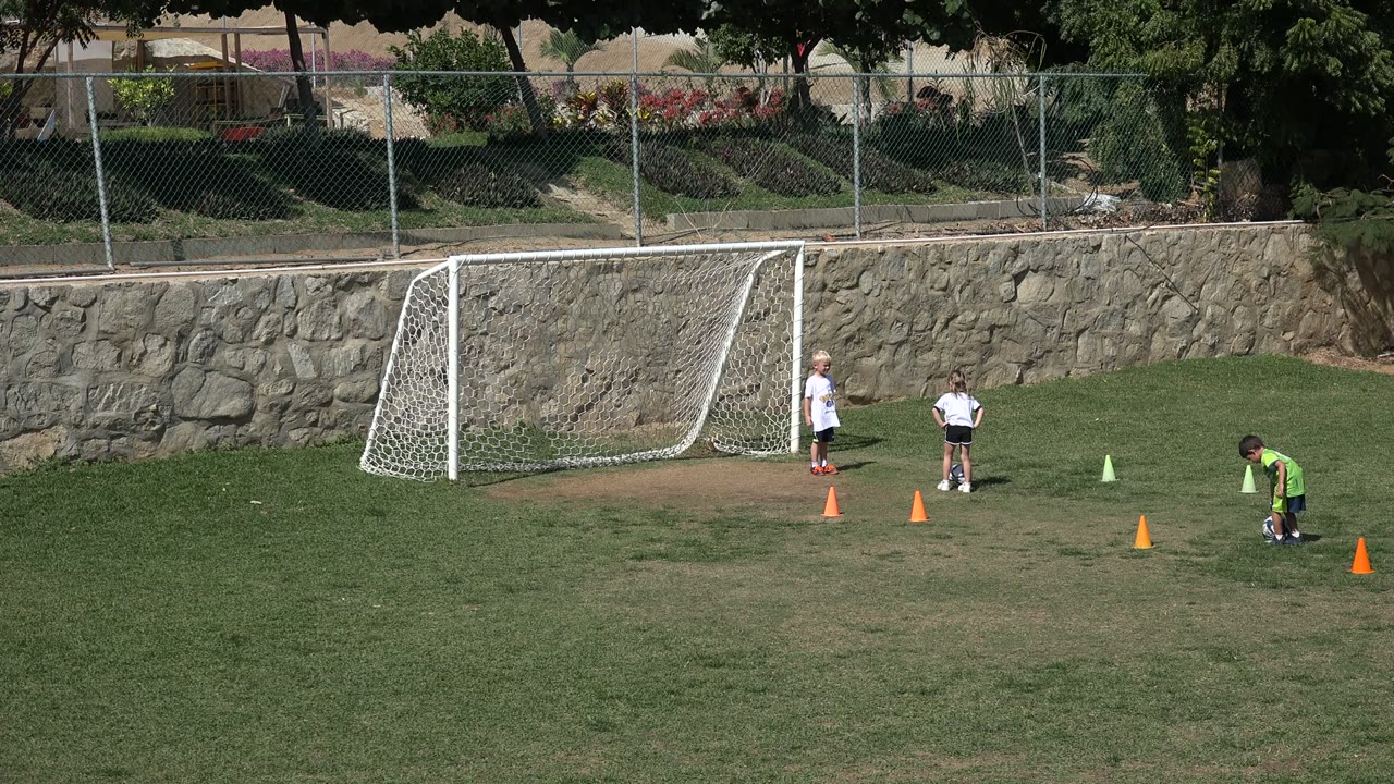 Children stand and move near a small soccer goal on a grass field in Los Cabos, Mexico, with orange cones and a stone wall behind them.