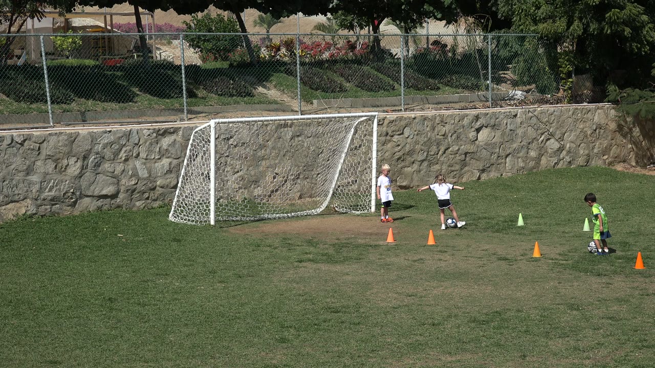 A child in a white shirt kicks a soccer ball between orange cones near a small goal in Los Cabos, Mexico, while other children stand nearby.