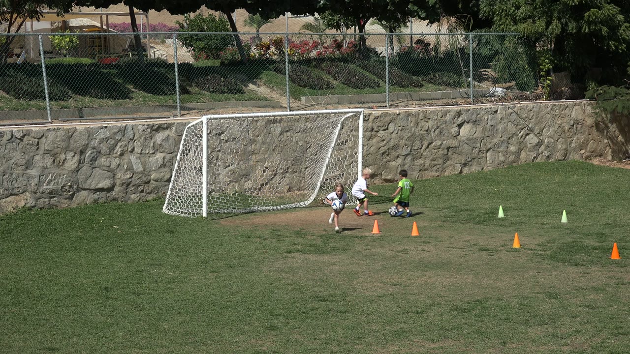 Three children gather near soccer balls and orange cones beside the white goal on a Los Cabos grass field, with a stone retaining wall behind them.
