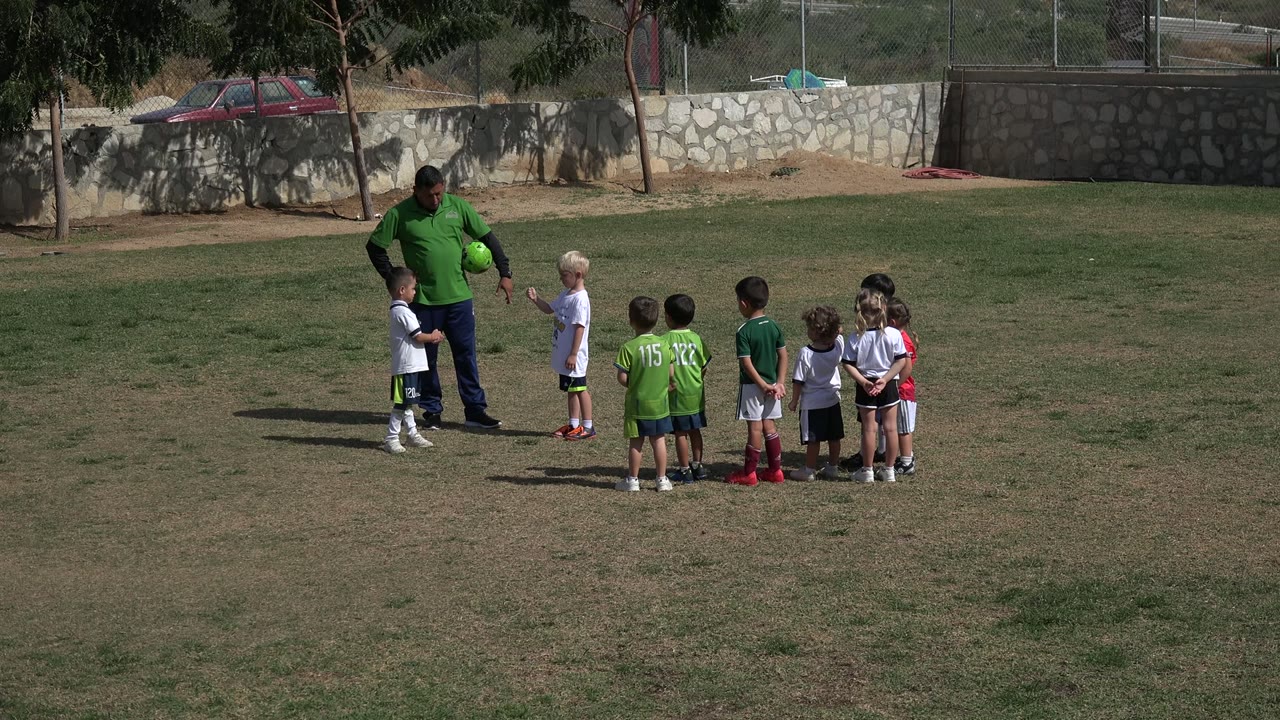 In Los Cabos, Mexico, the adult holds a soccer ball while children in white and green jerseys stand facing him on the grass.