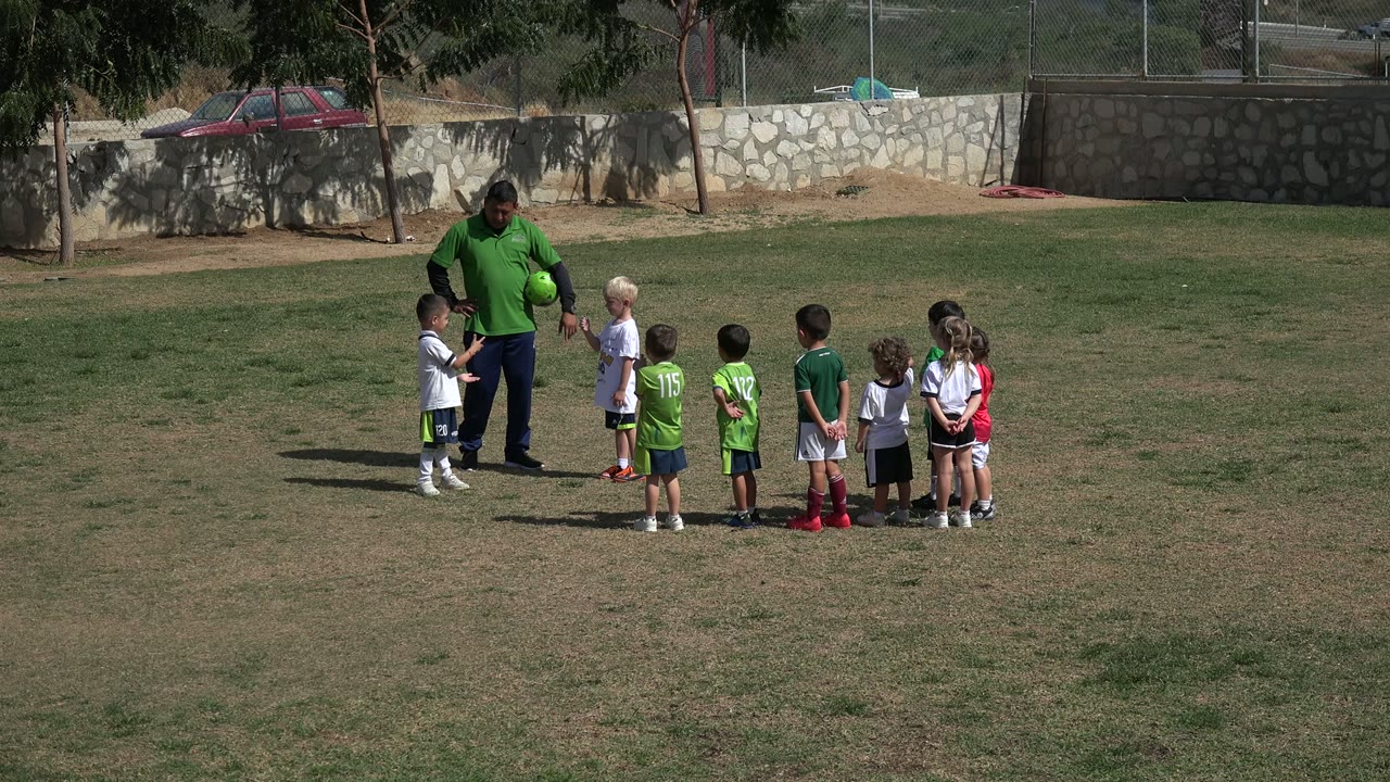 The adult remains near the center of the group as children in green and white sports clothes wait on the grass field in Los Cabos, Mexico.