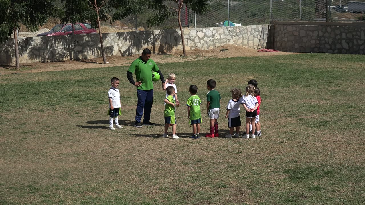 In Los Cabos, Mexico, a coach in a green shirt holds a bright green soccer ball while several young soccer players stand in a loose line on the grass.
