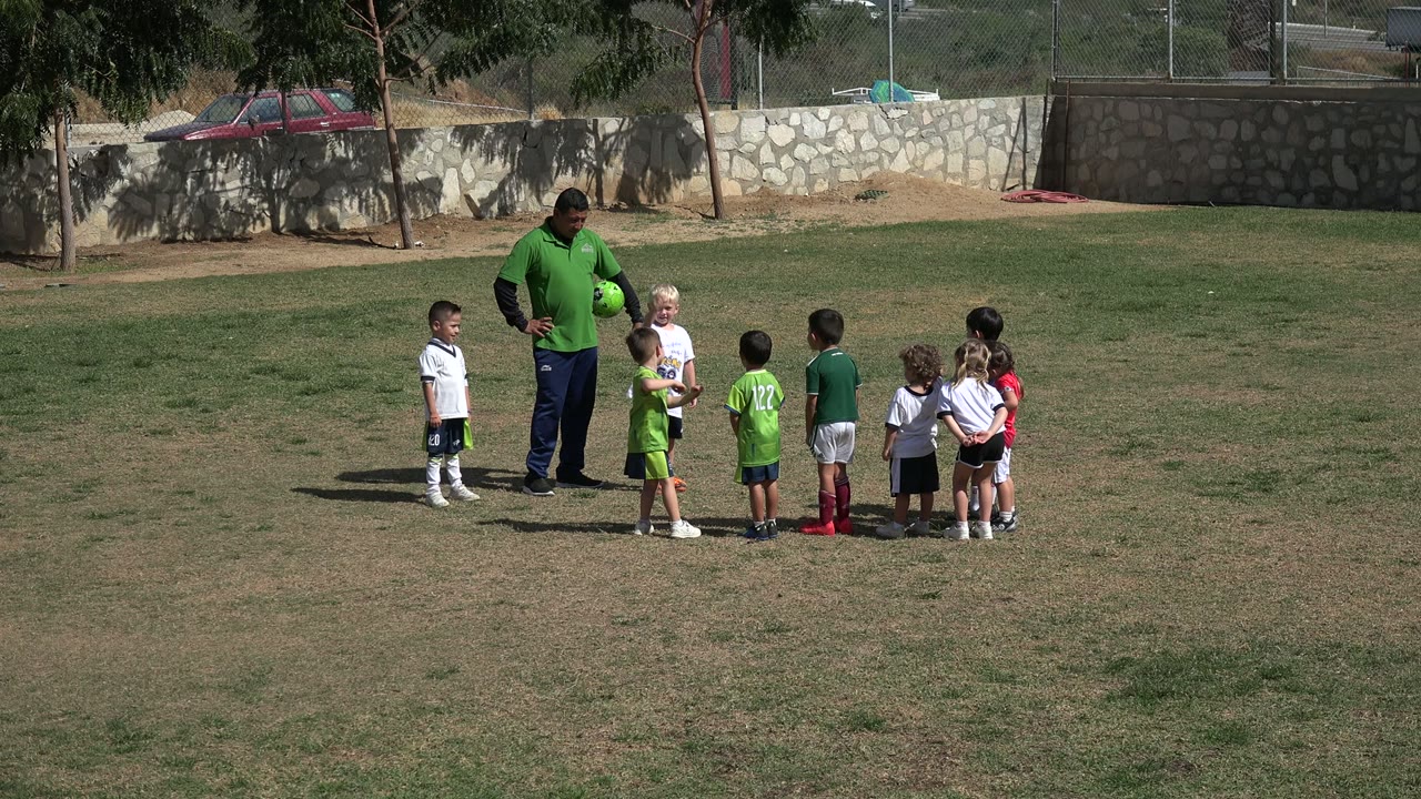 At one second, the coach remains near the group as children in white and green soccer uniforms stand on the dry grass in Los Cabos, Mexico.