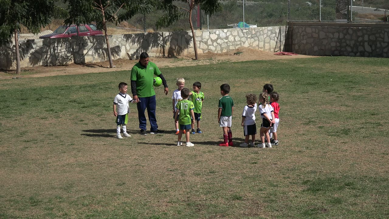 In Los Cabos, Mexico, children in white and green soccer outfits face a man holding a green ball, with open grass stretching around the group.