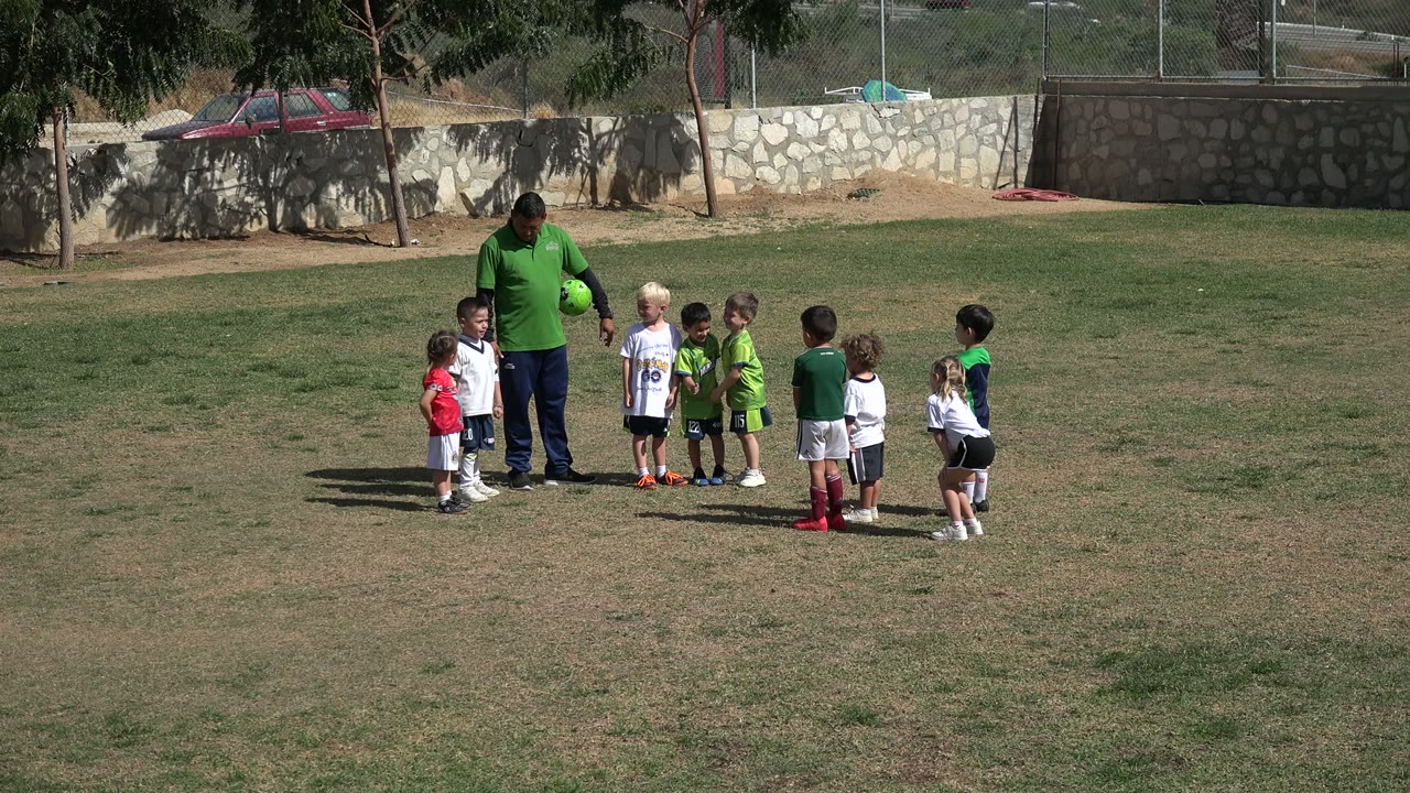 The adult remains centered with the ball while children in red, white, green, and dark sportswear face him on the Los Cabos grass field.