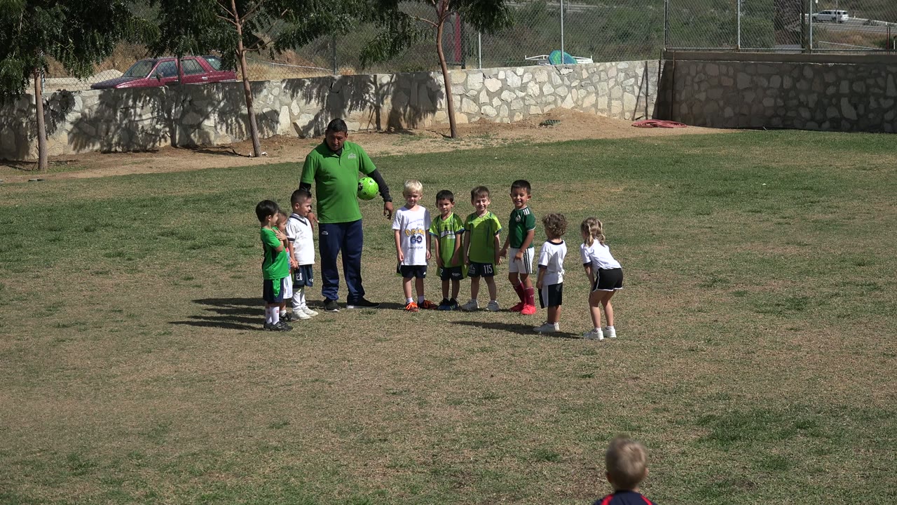 A group of children stands in a loose line near an adult holding a green ball on a Los Cabos grass field, with a wall, fence, trees, and parked car in the background.