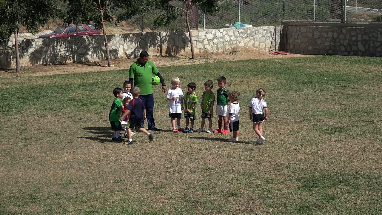 In Los Cabos, Mexico, several children shift positions around the coach as he holds a football on the field.