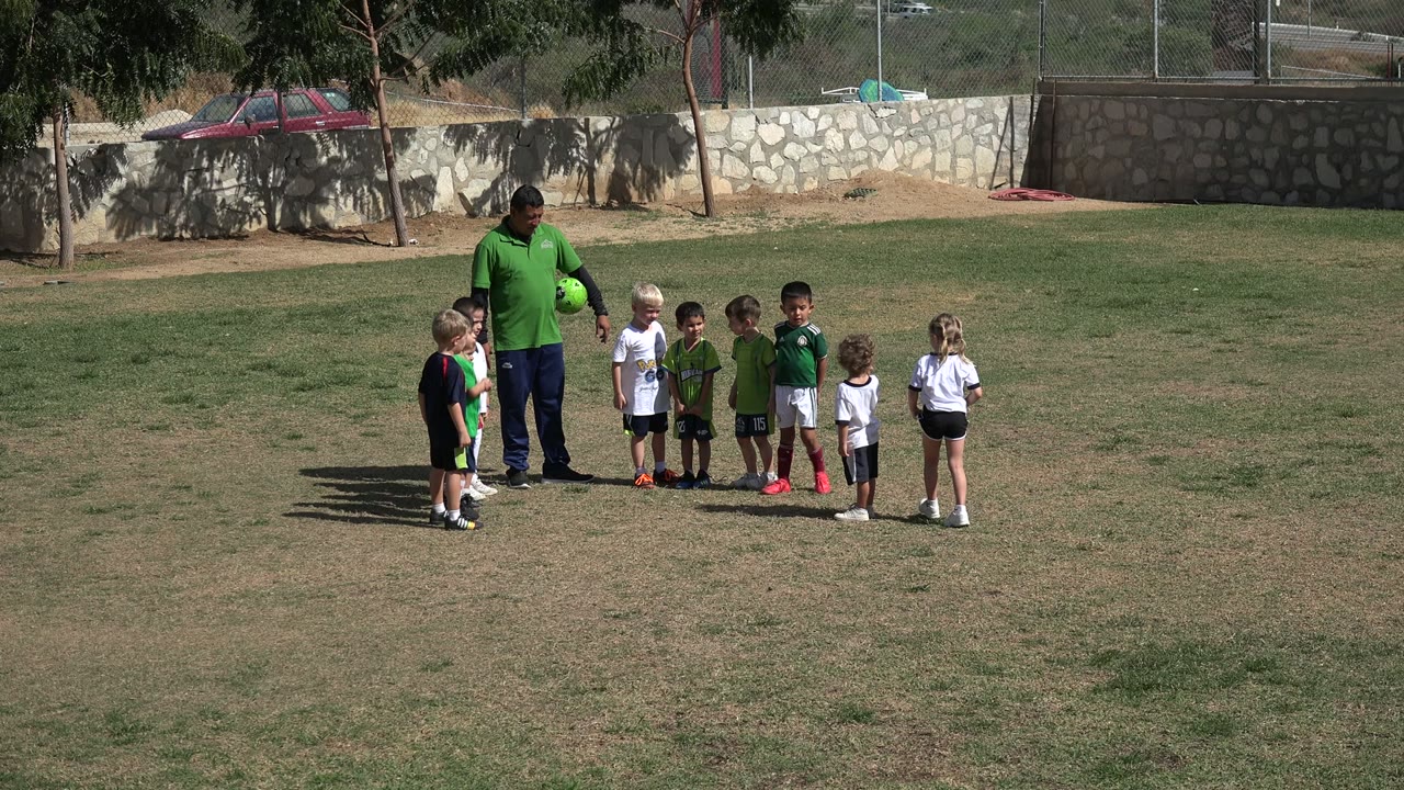 In Los Cabos, Mexico, a coach in a green shirt holds a soccer ball while children stand around him on a sunlit grass field.