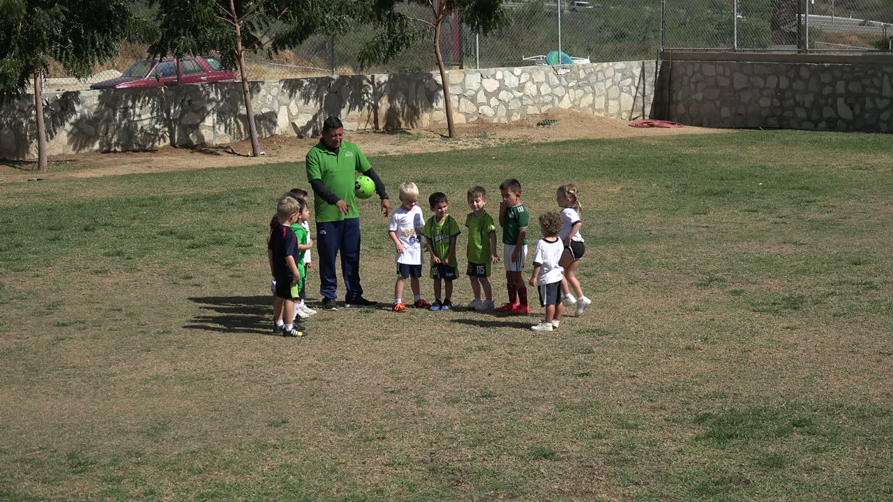 In Los Cabos, Mexico, several children in green, white, and dark sports shirts face a coach holding a ball on the grass.