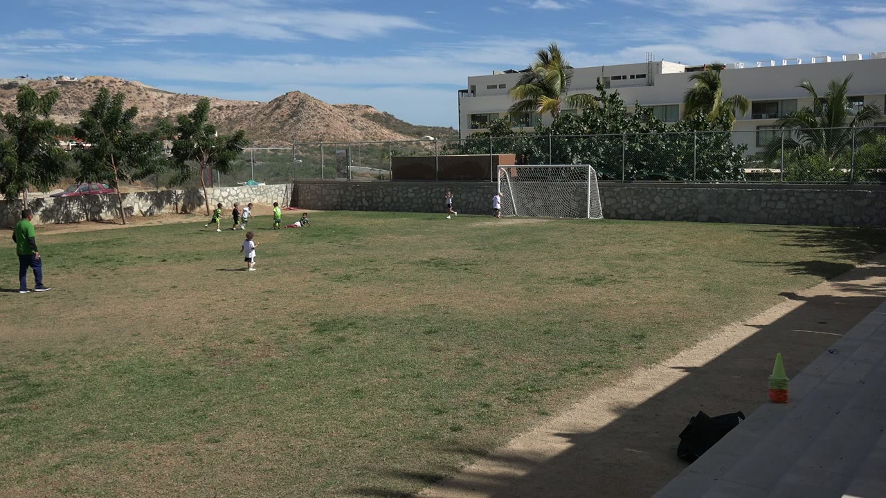 Children continue playing on the open grass field in Los Cabos, Mexico, with an adult standing on the sideline and the white goal, stone wall, palms, buildings, and hills behind them.