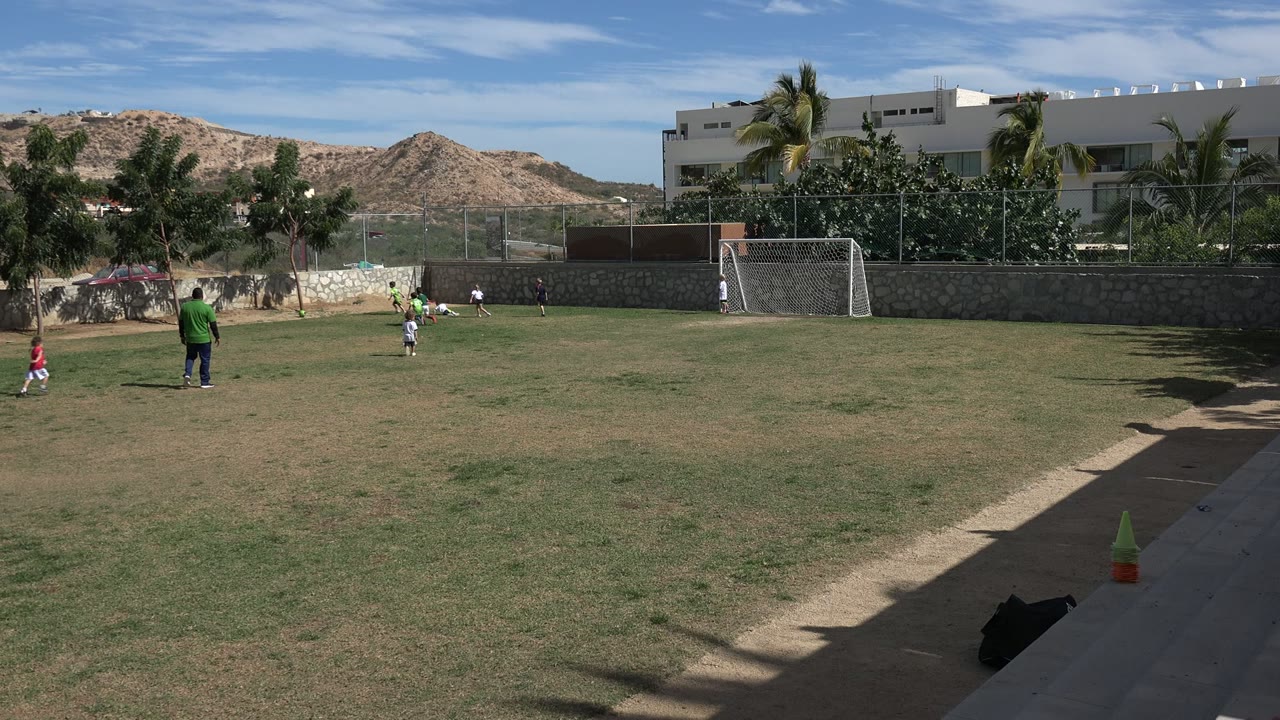 Children playing soccer on a grass field with a goal and hills behind them.