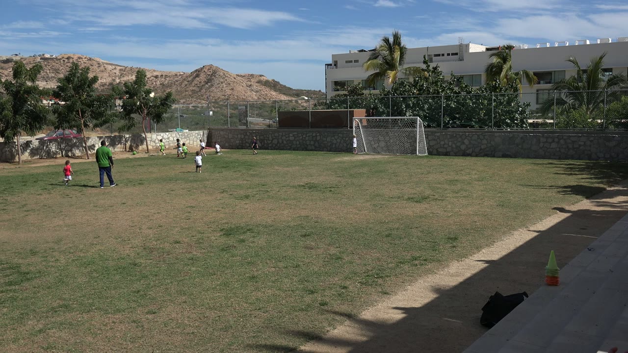 Children shift positions on the Los Cabos soccer field as an adult in a green shirt stands nearby, with the goal and shaded walkway still framing the right side.