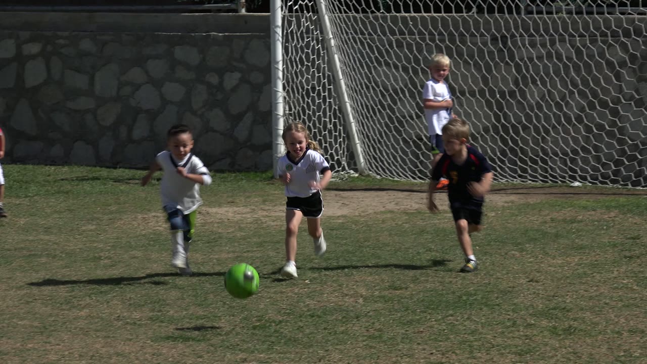 A group of young players run after a green soccer ball on a patchy field in Los Cabos, Mexico, with the goal net partly visible behind them.