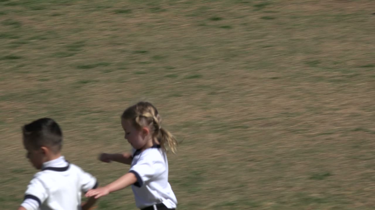 A girl in a white shirt and a boy run near a white soccer ball on a patchy grass field in Los Cabos, Mexico.