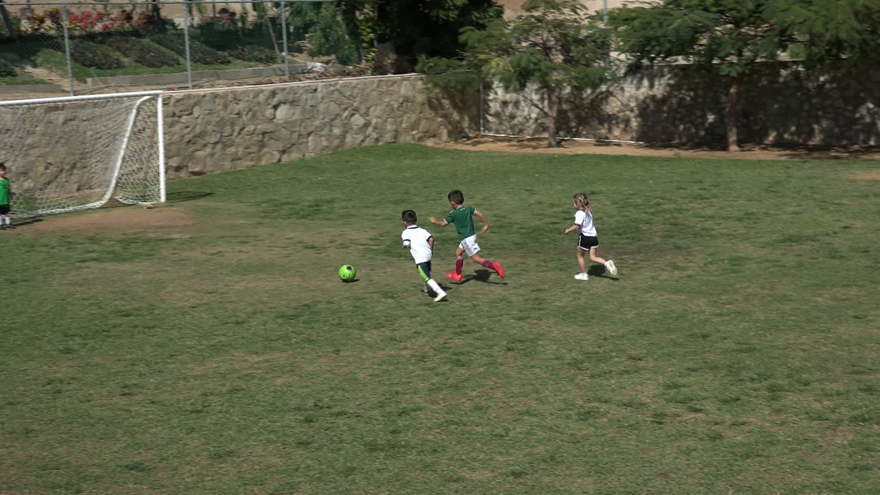 A child in green and a child in white run toward a green soccer ball on the Los Cabos field while another child stands near the white goal.