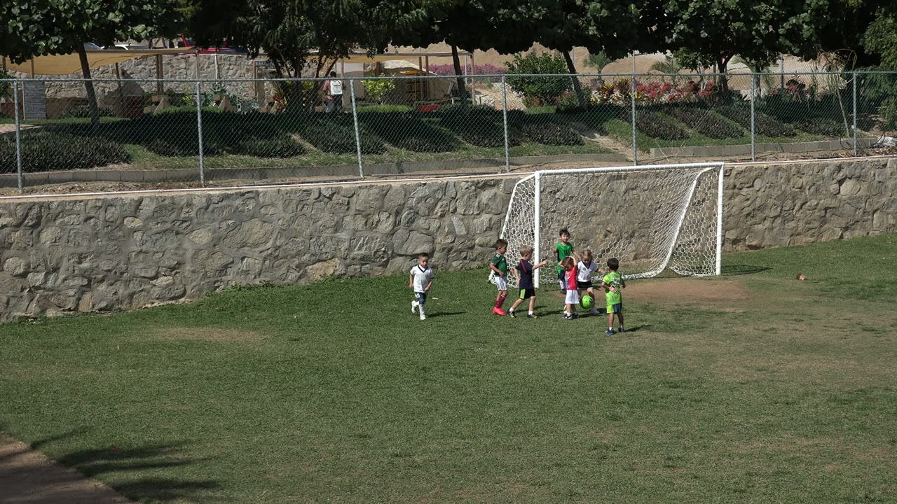 Children in soccer uniforms cluster near a small white goal on a sunny grass field in Los Cabos, Mexico, with a stone wall and fence behind them.