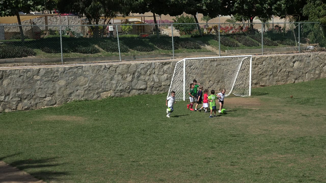 A group of children remains near the goalmouth on the Los Cabos soccer field, with one child in white standing apart and the stone wall running across the background.