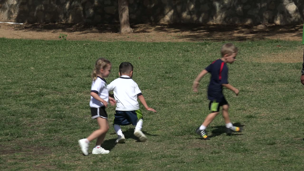 In Los Cabos, Mexico, small children in white and dark soccer outfits run across a grass field near the start of the clip.