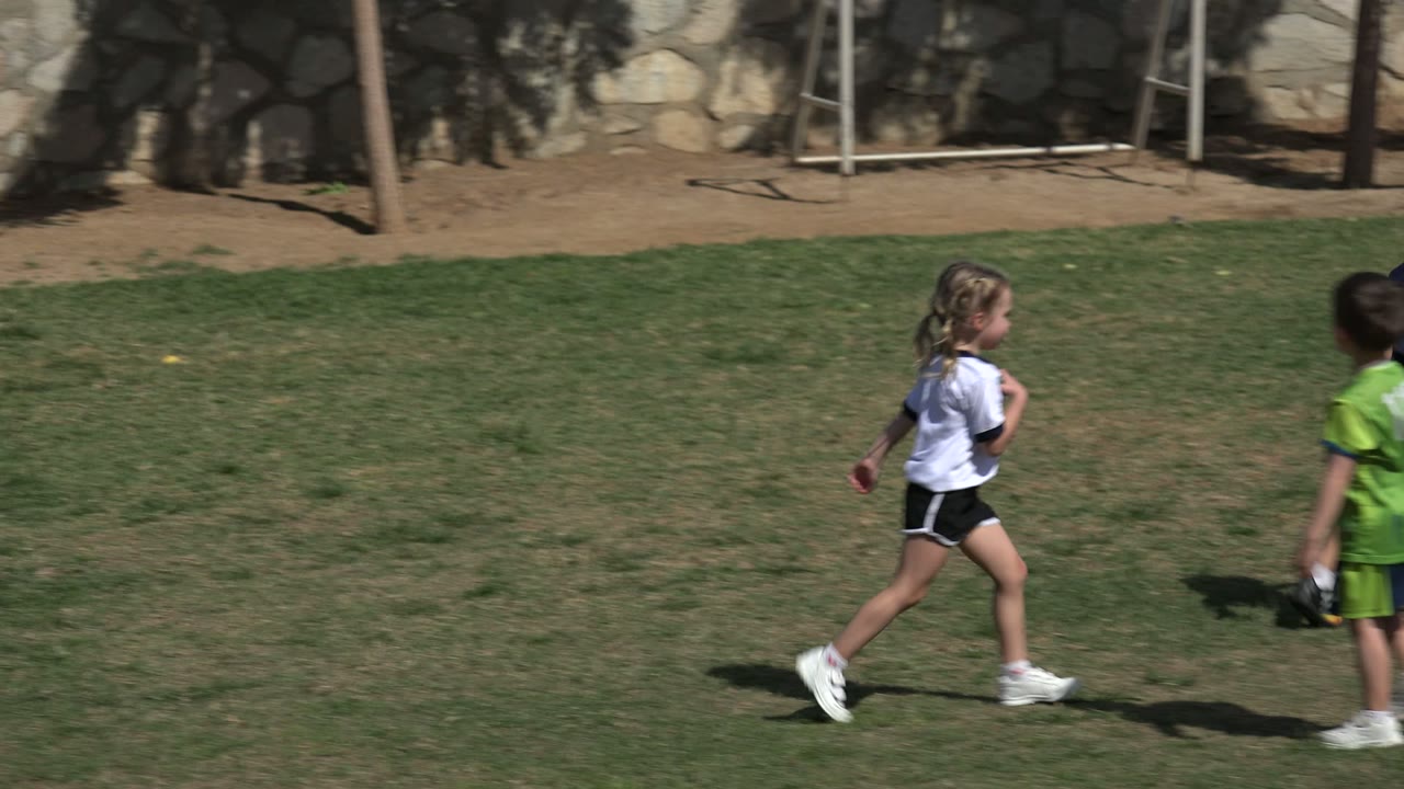 Several children cross the grassy field in Los Cabos, Mexico, while a bright green soccer ball sits near the right edge of the frame.