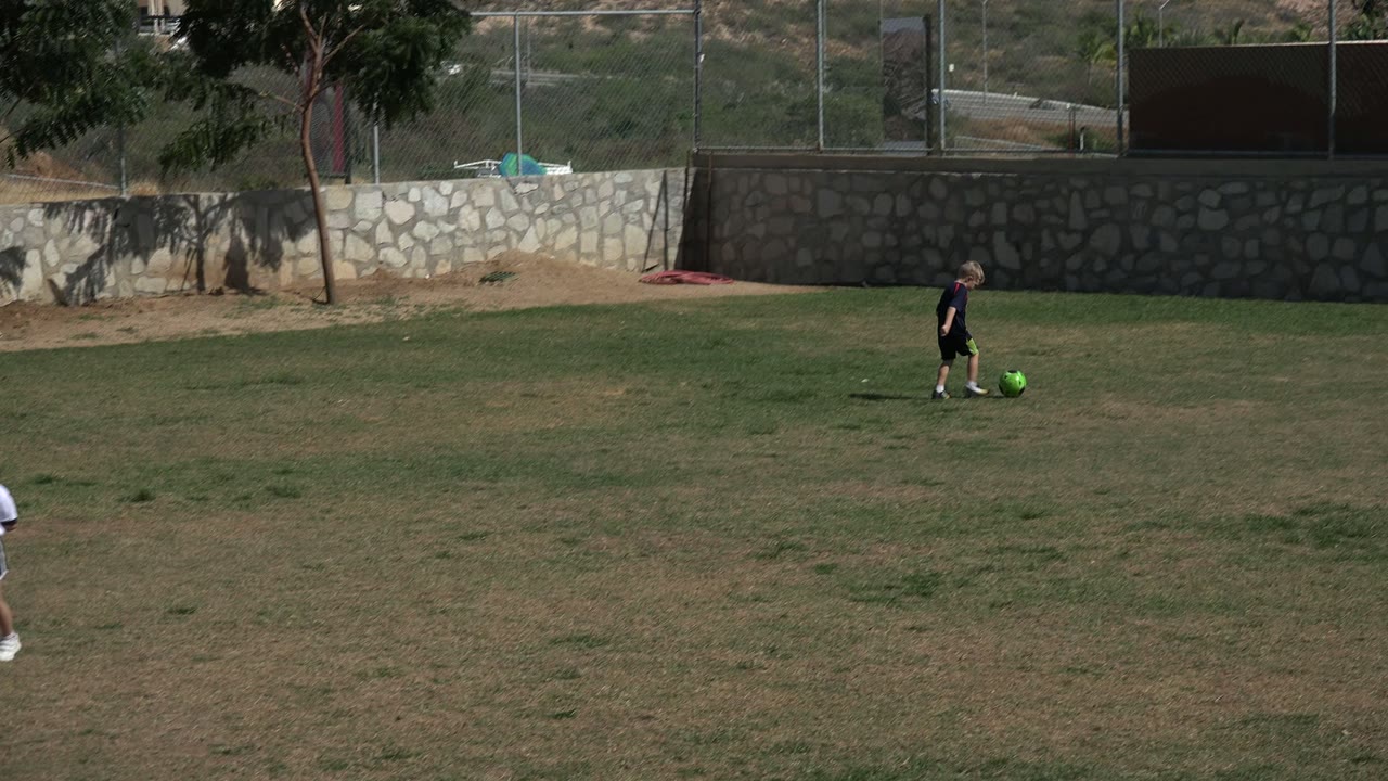 A child in dark clothing moves with a bright green soccer ball across the midground of a Los Cabos grass field, with a stone wall and fence behind.