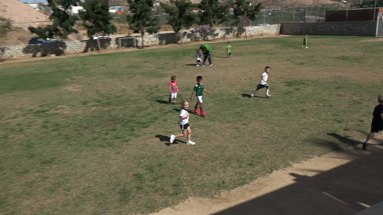 Children in white and green soccer uniforms move across the Los Cabos field as the green ball sits in the foreground, with trees, fencing, and dry hills in the distance.