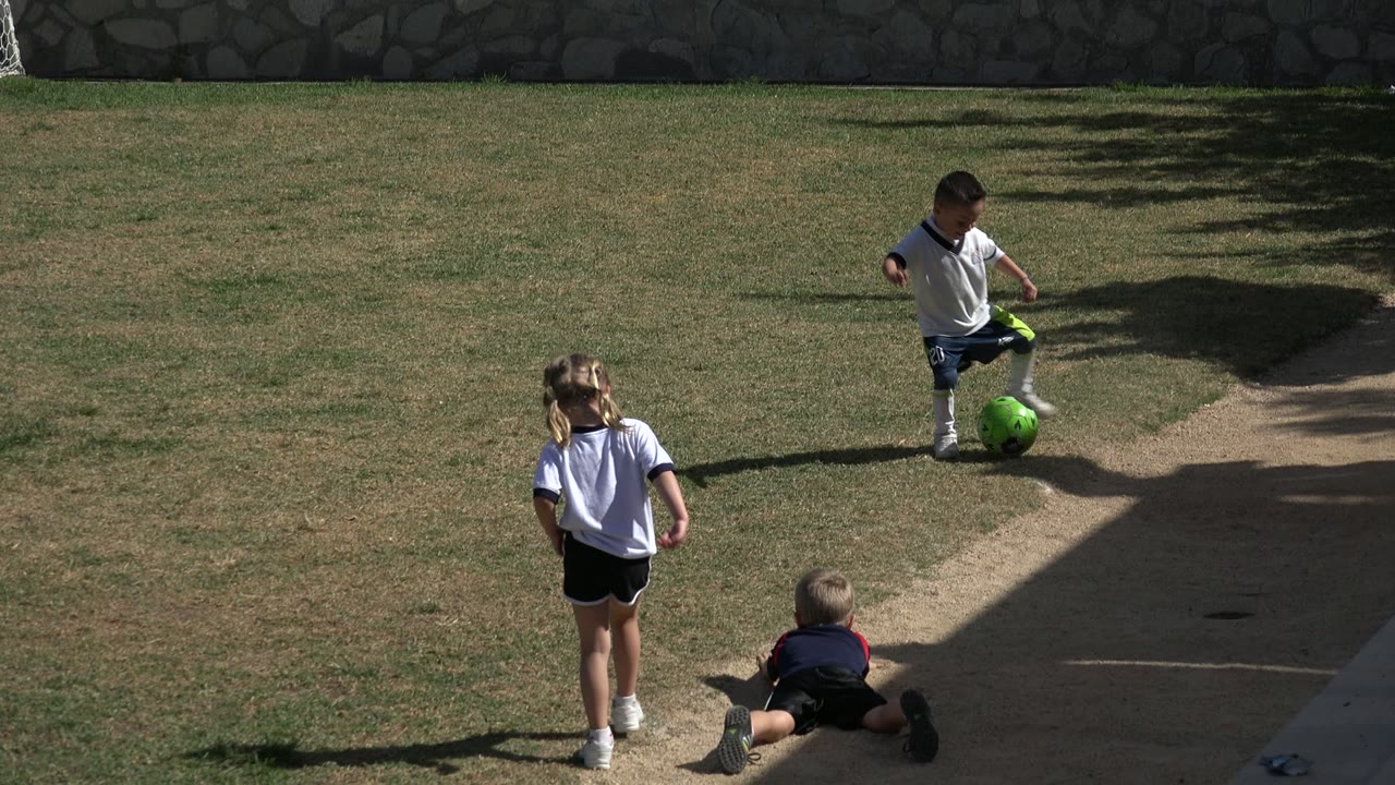 At the start of the Los Cabos soccer clip, children gather on a sunlit grass field as one boy controls a bright green ball near a concrete walkway.