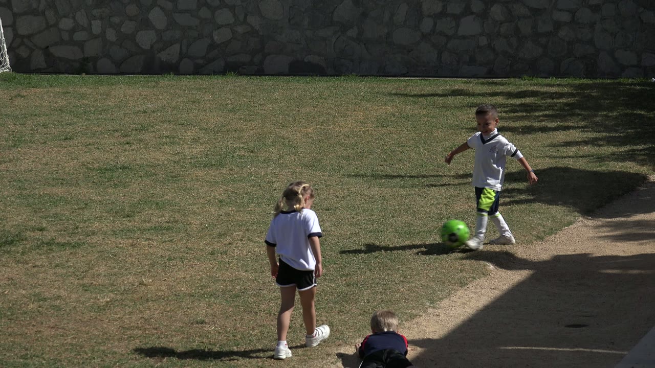 A boy in soccer clothes moves beside the bright green ball in Los Cabos while two other children remain nearby on the sunny grass field.