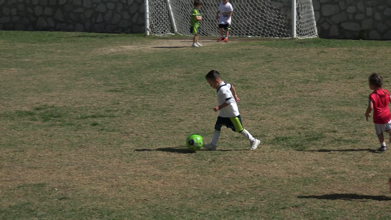 At 0:00 in Los Cabos, Mexico, a small boy in a white soccer shirt dribbles a bright green ball across patchy grass while another child in red and goal nets sit behind him.