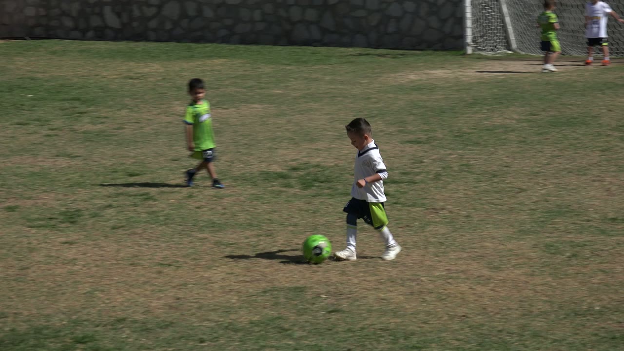 At 0:01 in Los Cabos, Mexico, the boy in white follows the green soccer ball across the field while an adult in a green shirt and other children appear near the goals.