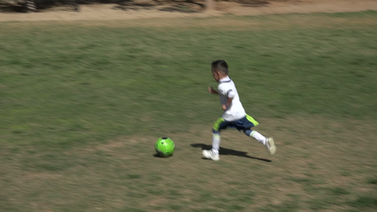 Young boy running after a bright green soccer ball on a grass field.